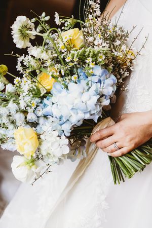 Bride holding lush wedding bouquet of blue hydrangeas, yellow roses, white daisies, and wildflowers with diamond ring.