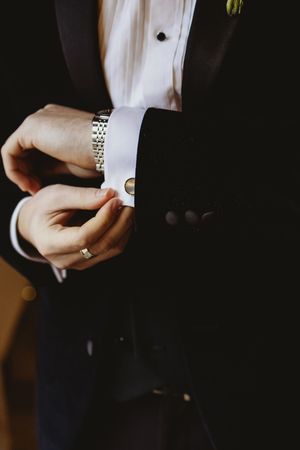 Groom in black tuxedo fastening silver cufflinks on white dress shirt with gold wedding band and watch.