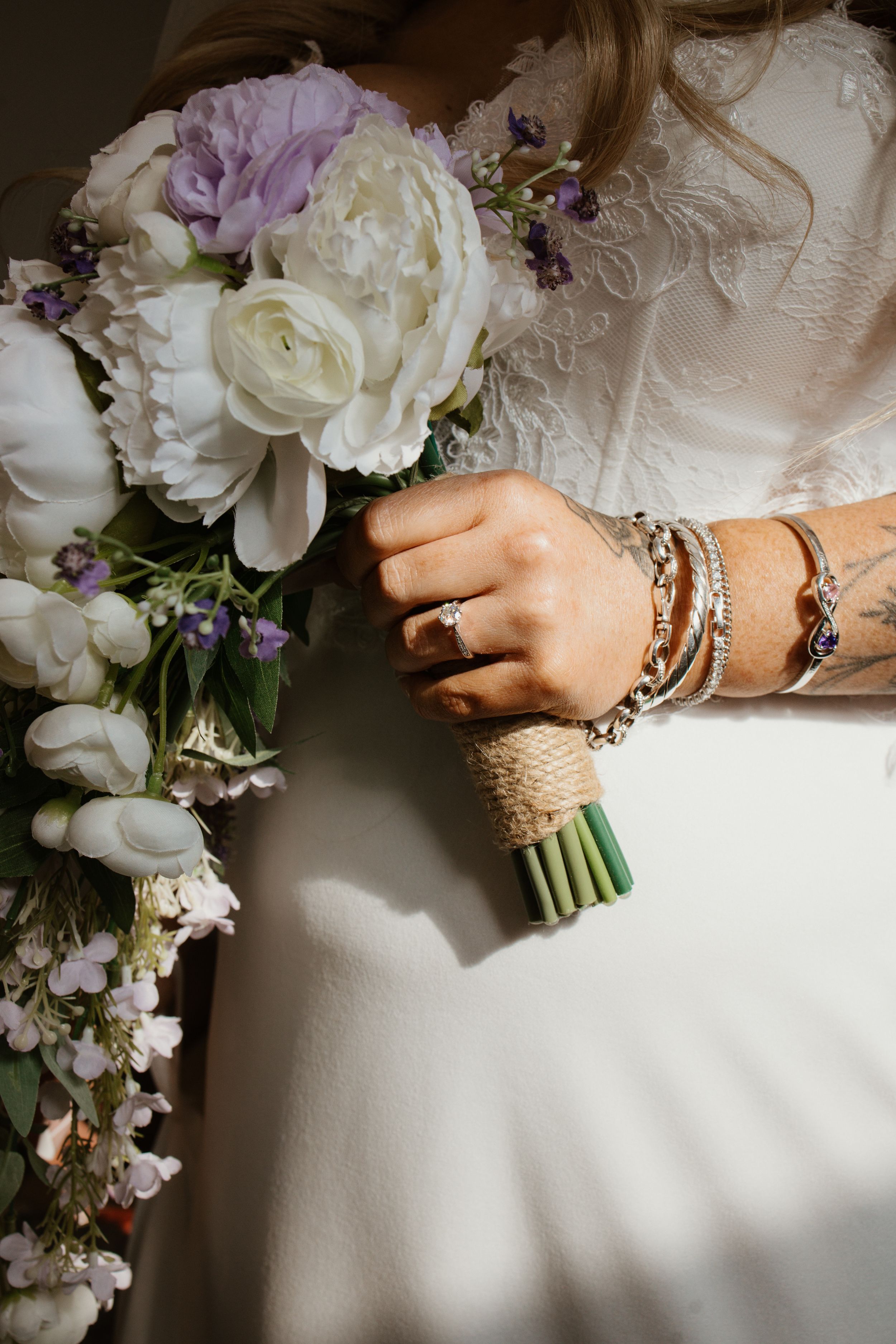Bride holding a white and lavender wedding bouquet wrapped in burlap, wearing a diamond ring and stacked bracelets.