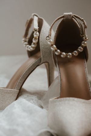 Close-up of elegant bridal heels adorned with pearl ankle straps and gold details on white lace fabric.