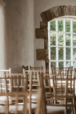 Rustic wedding ceremony venue with wooden chiavari chairs and arched stone window with garden view.