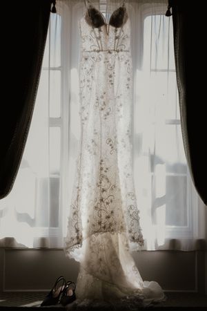 Intricate lace wedding dress hanging in window light with black heels placed below.