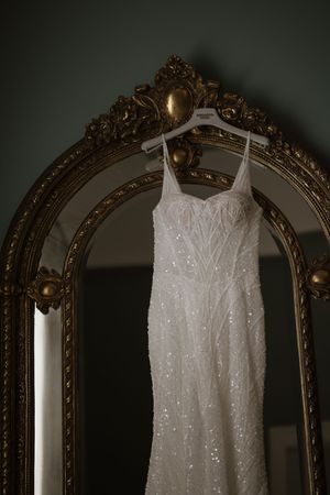 Beaded white wedding dress hanging on ornate gold vintage mirror against dark green wall.