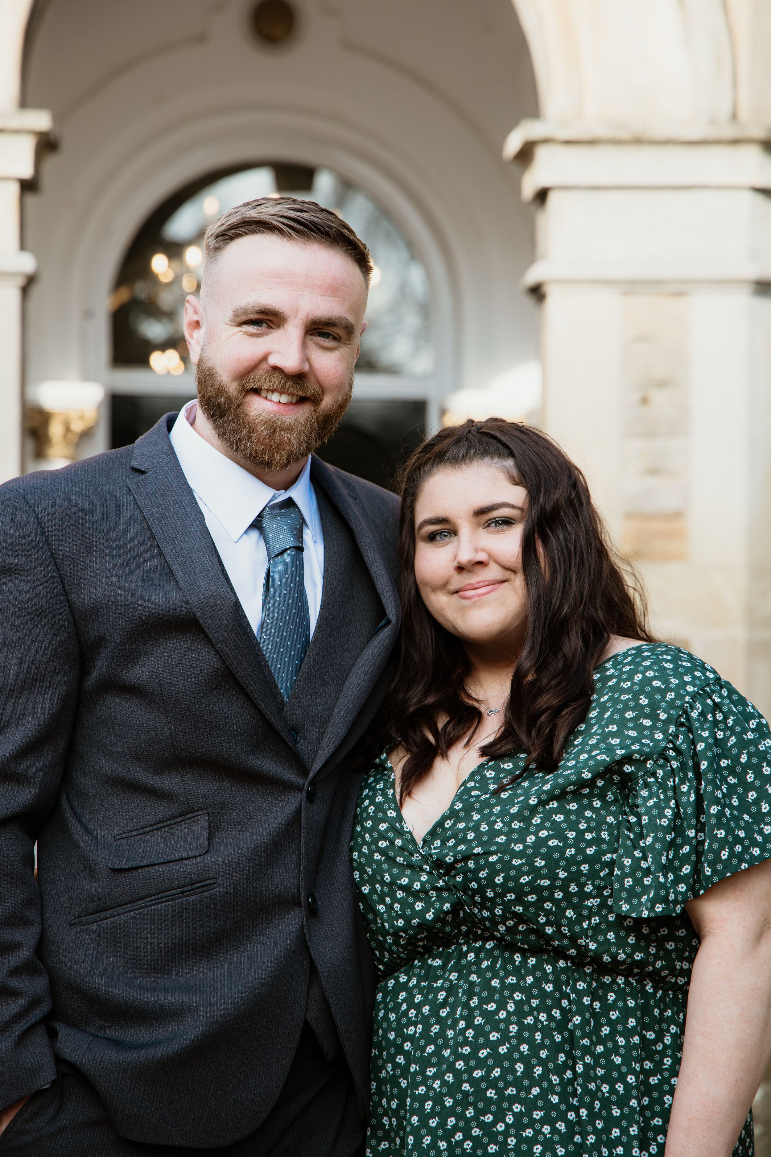 Smiling couple posing together outside a stone building, man in grey suit and woman in green floral dress.