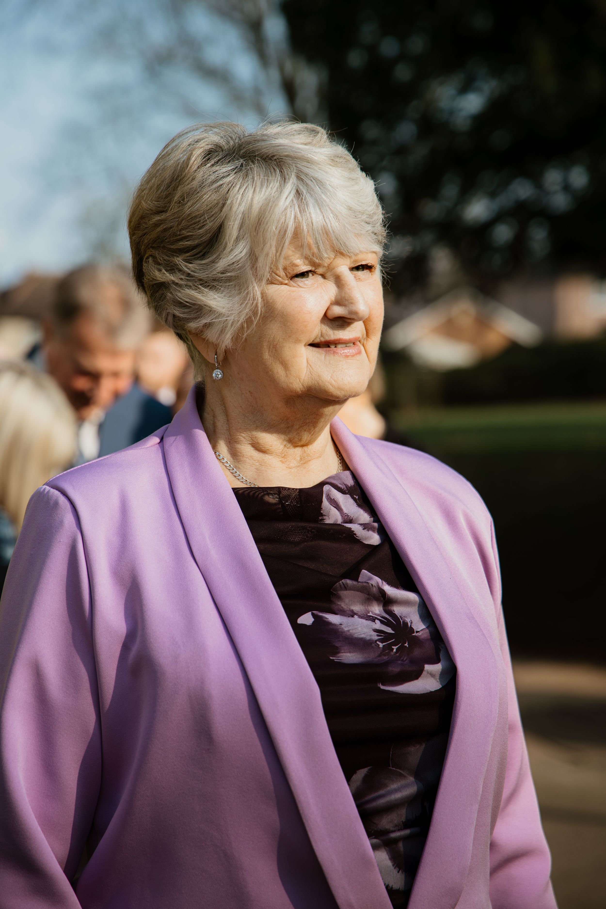 Elegant woman in a lavender blazer and floral dress smiling outdoors at a wedding event.
