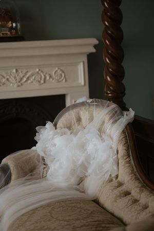 Ruffled white bridal veil draped over antique tufted chair near fireplace in elegant room.