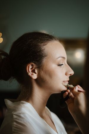 Bride in white robe receiving lipstick application during bridal makeup preparation session.