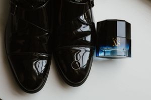 Black patent leather dress shoes beside a cologne bottle on white surface.
