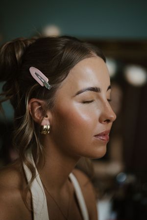 Bridesmaid with gold hoop earrings and pink hair clip showing glowing makeup during bridal preparations.
