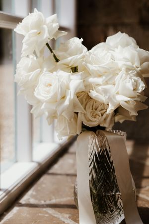 Elegant white roses bouquet in a silver vase with ribbon by a window.