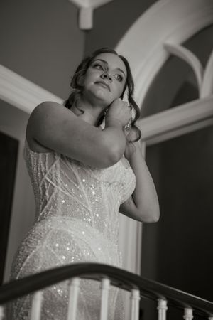 Bride in a sequined strapless wedding gown adjusting her earring on a staircase in a black and white photo.