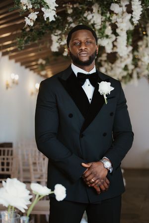 Groom in a black double-breasted tuxedo with white boutonniere standing under floral arch at wedding.