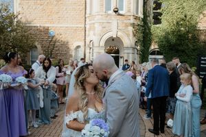 Newlyweds sharing a kiss outside a stone church surrounded by wedding guests in purple attire.