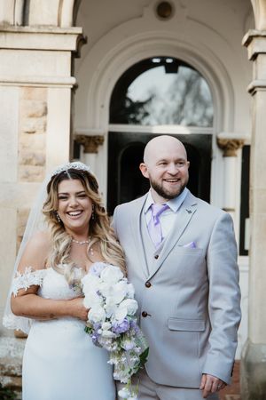 Laughing bride in tiara and lace gown with groom in light grey suit holding white wedding bouquet.