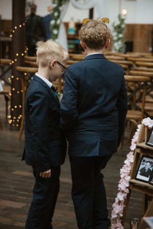 Two young boys in navy suits walking down wedding ceremony aisle from behind.