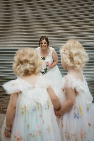 Smiling bride in lace gown holding bouquet facing two blonde flower girls in floral dresses outdoors.