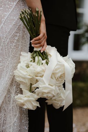 Bride holding a lush bouquet of white roses tied with white satin ribbon at an outdoor wedding.