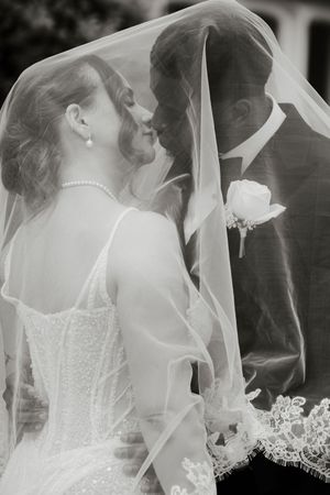 Black and white photo of bride and groom leaning close together under a lace wedding veil.