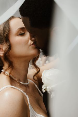 Bride and groom sharing a romantic kiss under a flowing white veil on their wedding day.