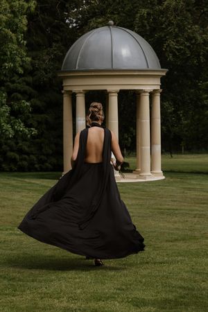 Bridesmaid in a flowing black backless gown walking toward a stone gazebo in a lush garden.