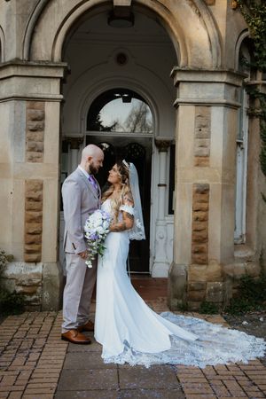 Bride and groom gazing at each other under a stone archway, bride's lace veil trailing on ground.