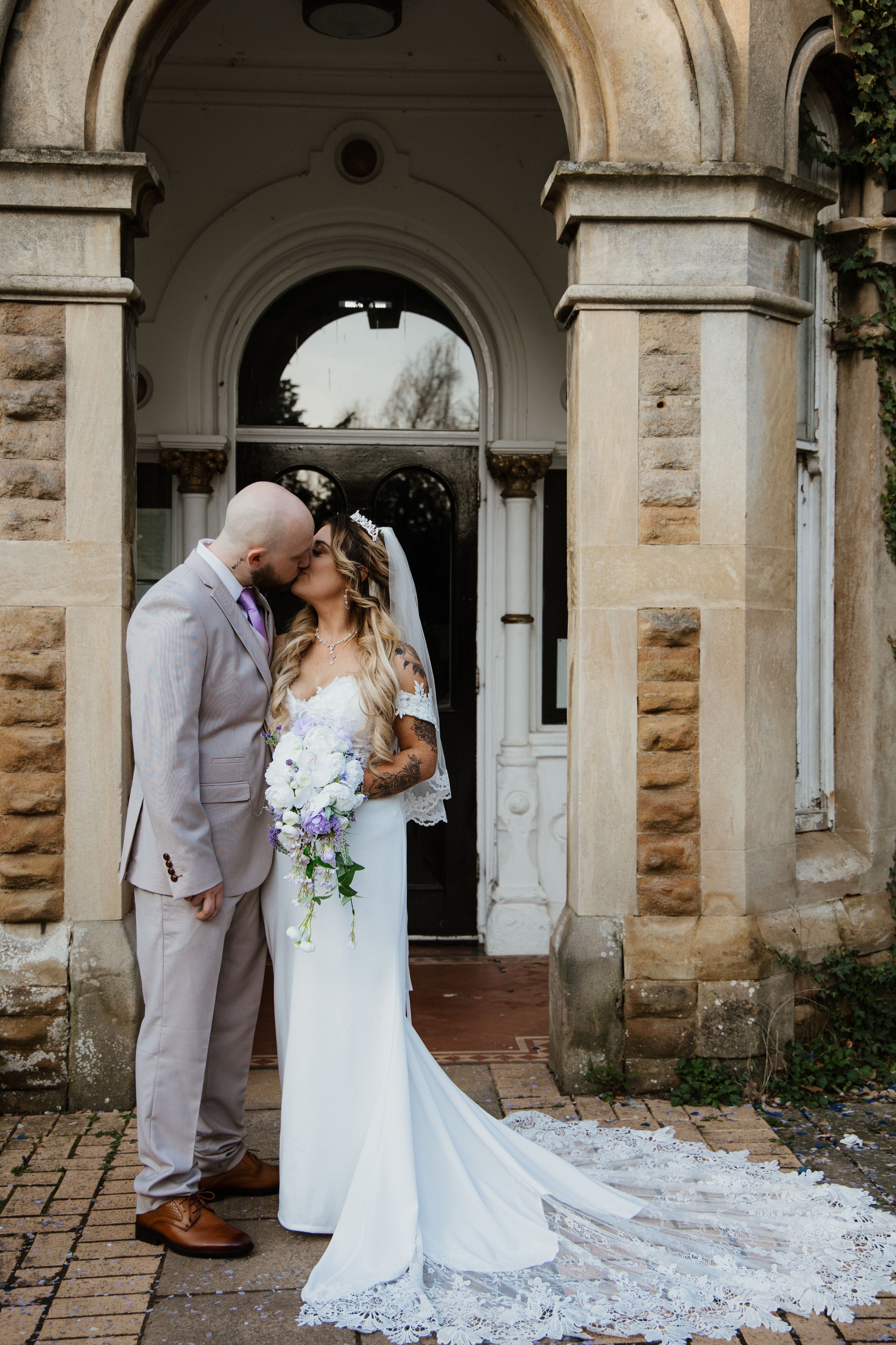 Bride and groom sharing a kiss under a stone archway, bride in lace gown with cathedral veil.