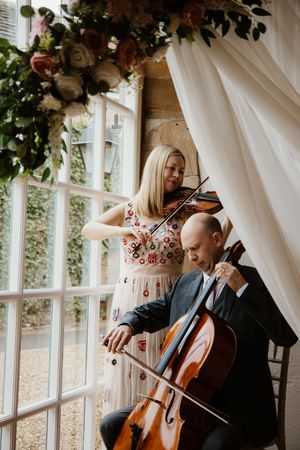 Violinist and cellist duo performing at wedding ceremony beside floral arch and large window.