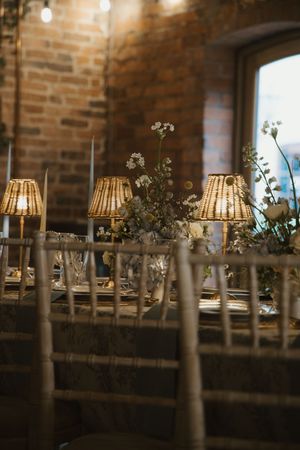 Rustic barn wedding reception table with rattan lampshades, white candles, floral centerpieces, and exposed brick walls.