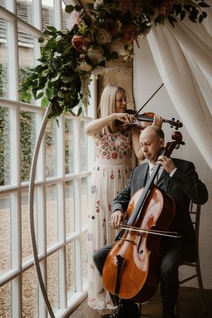 String duo playing violin and cello beneath floral arch at wedding venue.