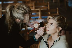 Professional makeup artist applying powder to bride with elegant updo hairstyle in rustic barn setting.