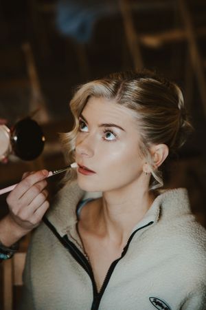 Blonde bride with elegant updo hairstyle getting makeup applied with brush in dimly lit barn venue.