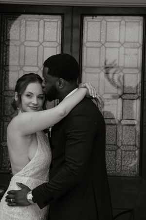 Black and white portrait of bride and groom embracing in front of stained glass windows.