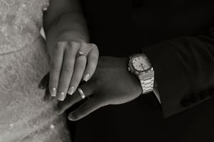 Close-up black and white photo of bride and groom hands showing wedding rings and watch.
