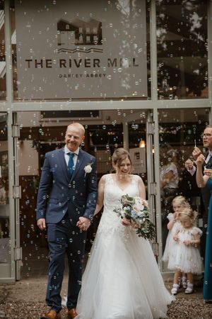 Newlyweds exit The River Mill Darley Abbey venue surrounded by bubbles with flower girls nearby.