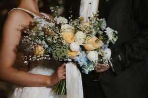 Bride and groom holding a lush wedding bouquet with peach roses, white ranunculus, and blue delphinium flowers.