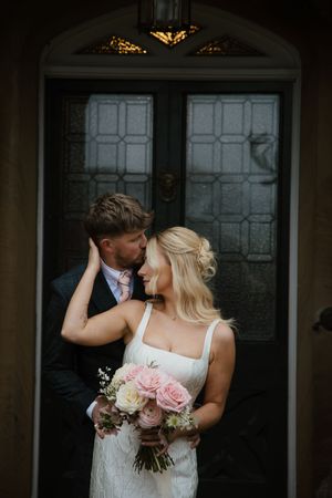 Groom tenderly kissing bride holding pink rose bouquet in front of ornate dark wooden doors.