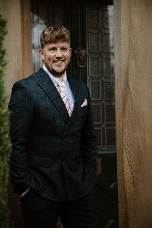 Smiling groom in dark plaid double-breasted suit with pink tie standing in doorway.