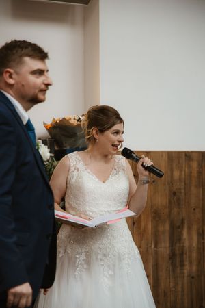 Bride in lace gown reading vows into microphone beside groom during wedding ceremony indoors.