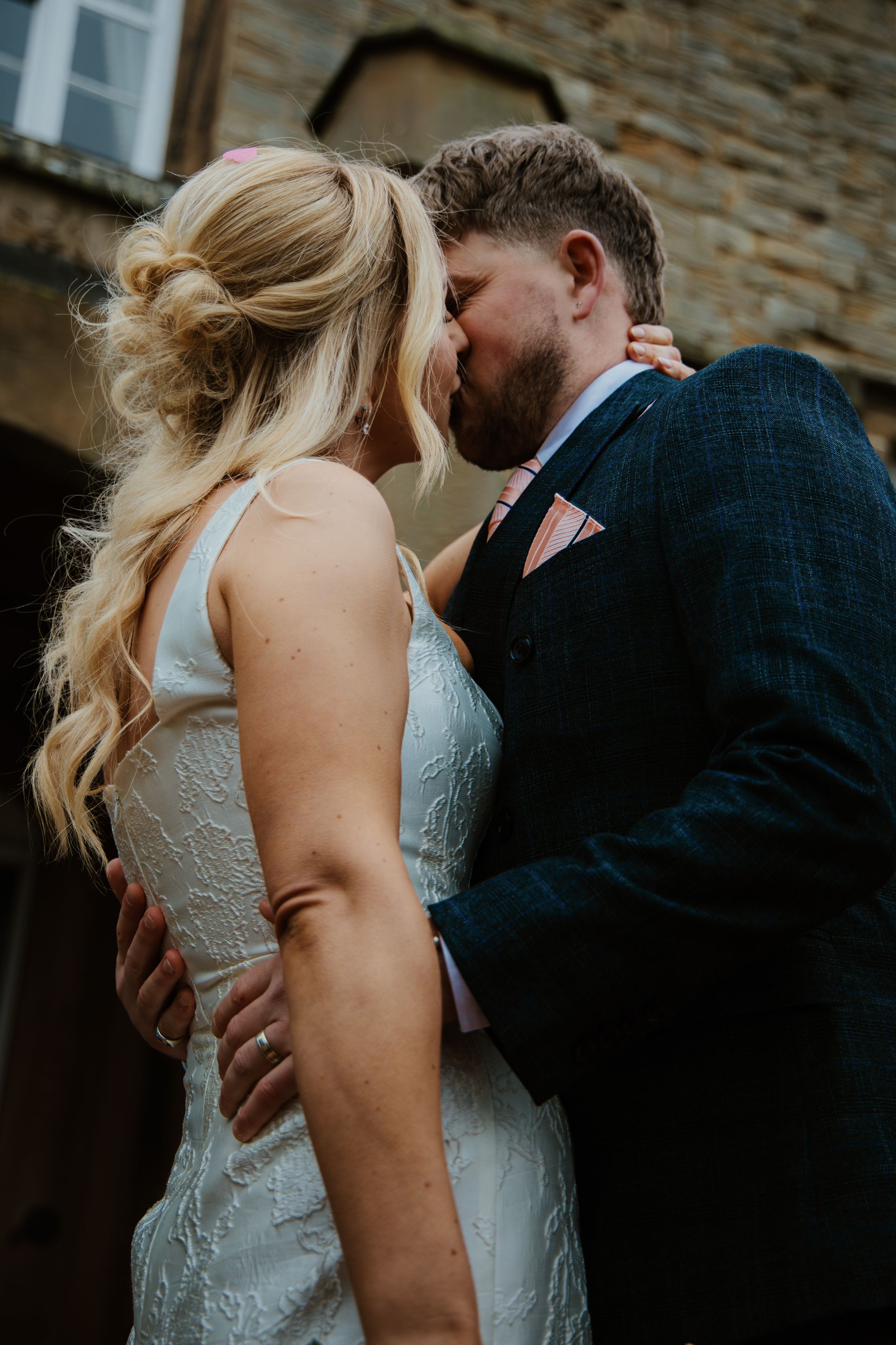 Newlyweds sharing first kiss outside rustic stone wedding venue in blue suit.