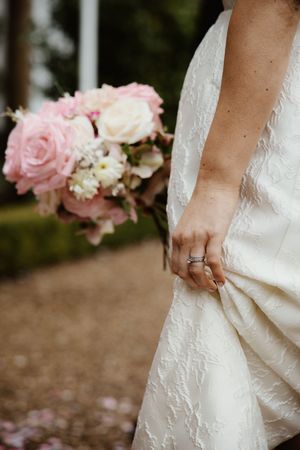 Close-up of bride holding pink rose bouquet with wedding rings visible on her hand.