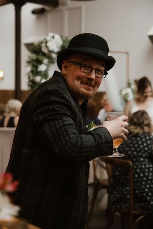 Smiling wedding guest in bowler hat and plaid jacket holding a beer at a wedding reception.