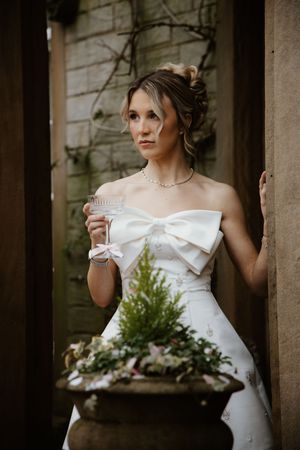 Elegant bride in strapless bow wedding gown holding champagne glass in rustic garden doorway.