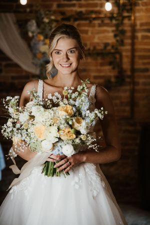 Smiling bride in white gown holding a beautiful bouquet of peach roses, white flowers, and blue blooms indoors.