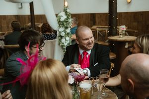 Man in black suit with red tie performing magic trick at wedding reception table with guests.