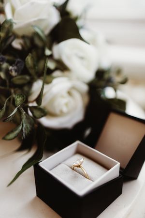 Gold diamond engagement ring displayed in black velvet box beside white roses bouquet.