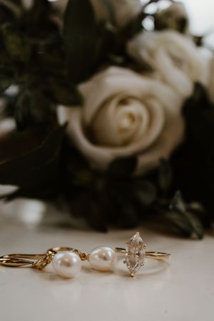 Gold marquise diamond ring and pearl drop earrings displayed beside white roses bouquet.