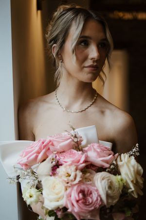 Elegant bride with updo hairstyle holding pink rose bouquet wearing pearl necklace.