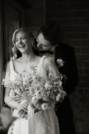 Black and white photo of laughing bride and groom embracing while holding a large floral bouquet.