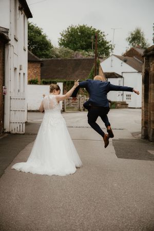 Joyful groom jumping in air while bride in white lace gown holds his hand on village street.