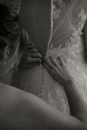 Black and white photo of hands buttoning intricate lace wedding dress from behind.